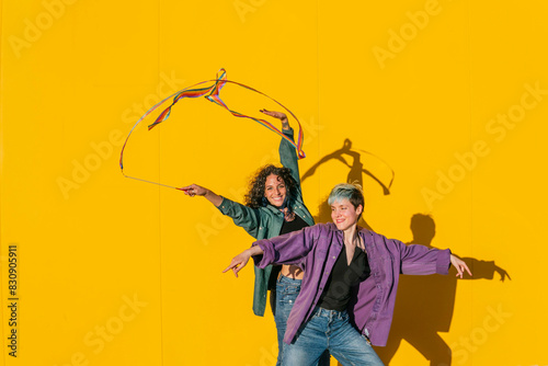 Smiling woman waving multi colored ribbon with lesbian friend in front of yellow wall photo