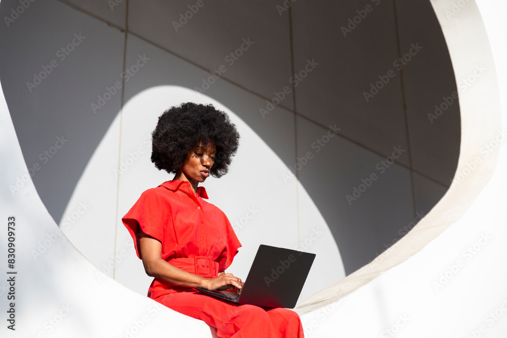 © VISTA by Westend61 - Woman in red jumpsuit with Afro hairstyle using laptop sitting on white wall © VISTA by Westend61 - Woman in red jumpsuit with Afro hairstyle using laptop sitting on white wall