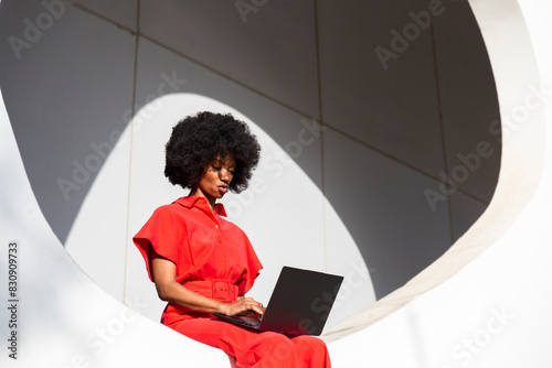 Woman in red jumpsuit with Afro hairstyle using laptop sitting on white wall