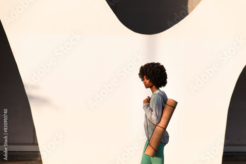 Woman with Afro hairstyle carrying exercise mat walking by white wall