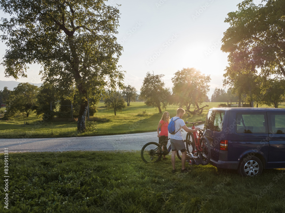 Young man is unloading electric mountain bikes, for himself and his girlfriend, lifting them from the hitch rack on the vehicle. Young man is unloading electric mountain bikes, for himself and his girlfriend, lifting them from the hitch rack on the vehicle.