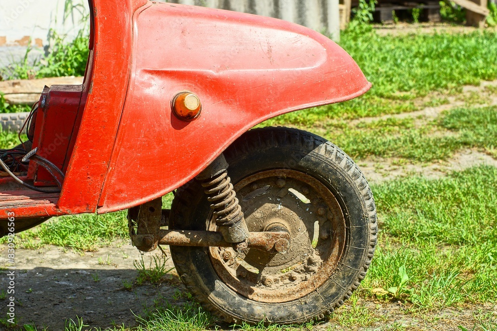 Fototapeta premium one round front dirty rusty iron wheel with a rubber black tire and an iron fender from a red retro scooter stands on the green grass during the day on the street