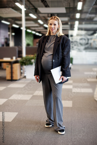 Confident pregnant businesswoman standing in a modern office, holding a tablet and smiling, showcasing balance between career and motherhood in a professional environment