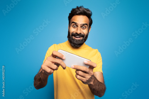 A joyful Indian man wearing a yellow shirt is enthusiastically playing a mobile game. He is set against a vibrant blue background, fully immersed in the game.