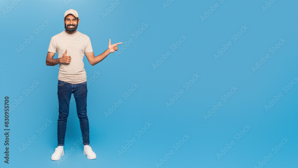 A casually dressed man stands against a vibrant blue backdrop, smiling while giving a thumbs up with one hand and pointing with the other, copy space A casually dressed man stands against a vibrant blue backdrop, smiling while giving a thumbs up with one hand and pointing with the other, copy space