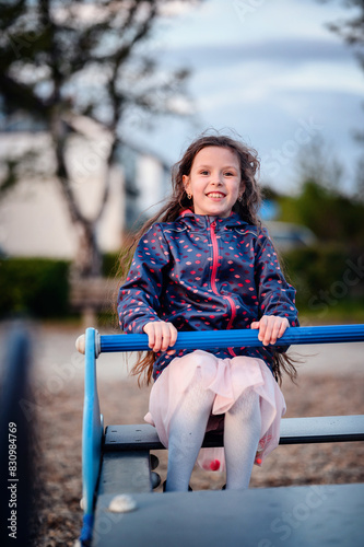 Happy young girl with long, wavy hair, wearing a blue jacket with pink patterns and a tutu, enjoying a ride on a seesaw in a park.