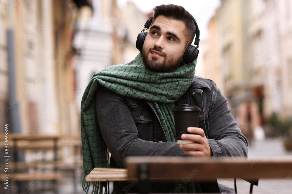 Fototapeta premium Smiling man in warm scarf with paper cup listening to music in outdoor cafe