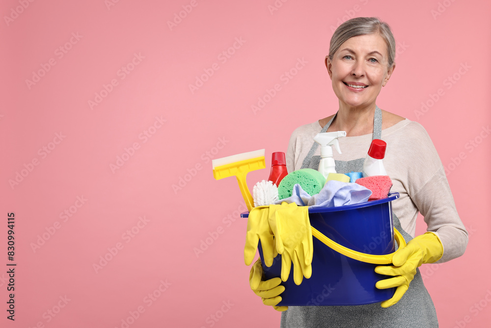 Happy housewife holding bucket with cleaning supplies on pink background, space for text