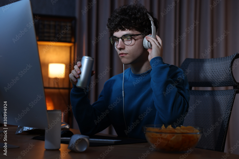 Young man with energy drink and headphones playing video game at wooden desk indoors Young man with energy drink and headphones playing video game at wooden desk indoors