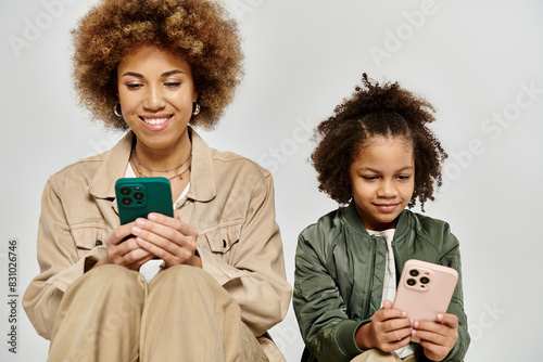 A curly African American mother and daughter in stylish clothes focus on their phones, engrossed in digital worlds.