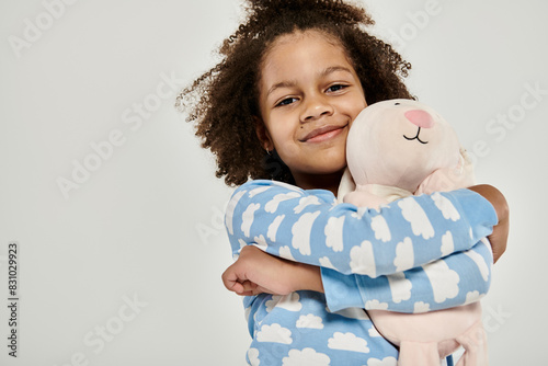 African American girl in pajamas cuddling a large stuffed animal on a grey background.