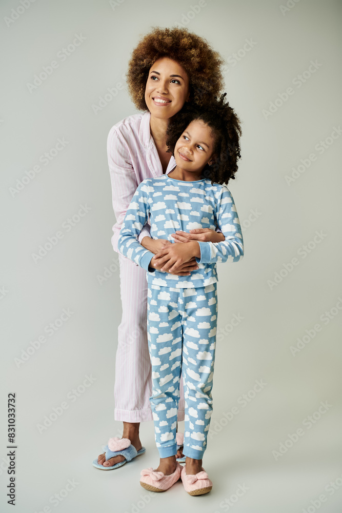 A cheerful African American mother and daughter strike a pose in matching pajamas against a grey backdrop. A cheerful African American mother and daughter strike a pose in matching pajamas against a grey backdrop.