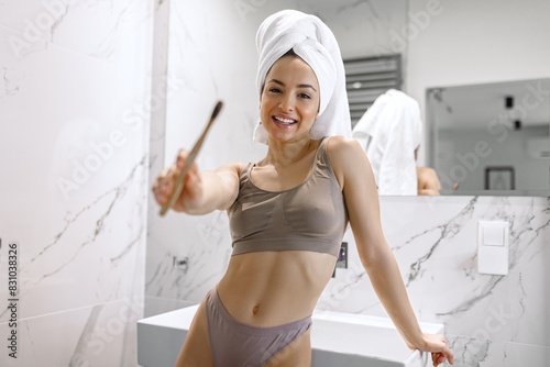 Satisfied young woman in underwear showing her toothbrush while standing near the washbasin with mirror in the bathroom. Lifestyle concept