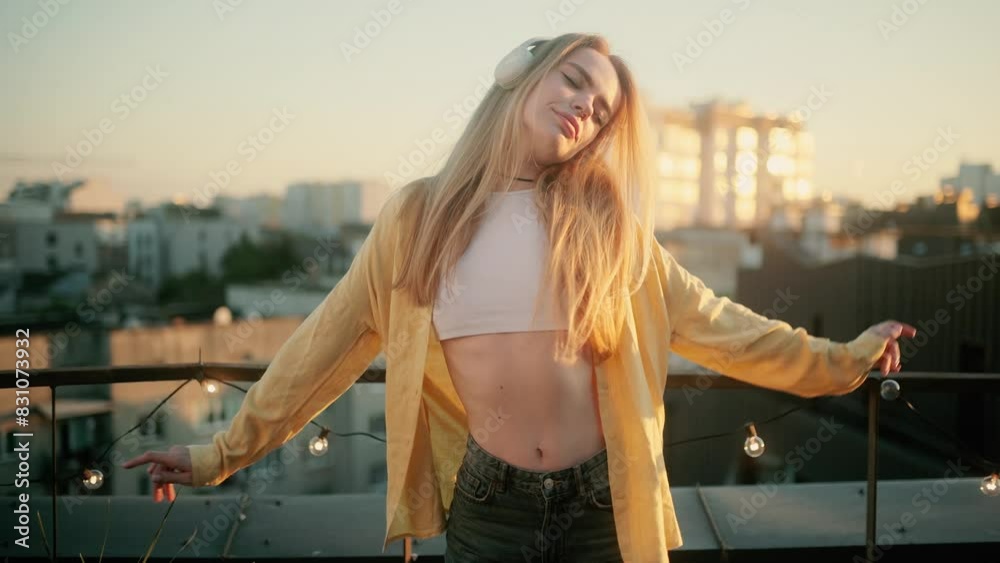 A young blonde woman in a white top and yellow shirt is dancing on a terrace during the sunset. A girl wearing headphones enjoying her favorite song A young blonde woman in a white top and yellow shirt is dancing on a terrace during the sunset. A girl wearing headphones enjoying her favorite song