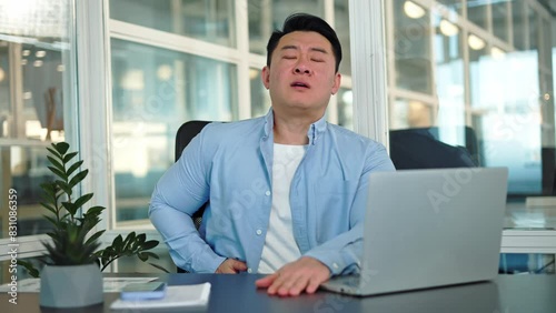 Portrait of regrettable man feeling inflammation of appendix during work process at personal laptop in office environment. Unhappy korean entrepreneur need medical help indoors. Concept of pain. photo