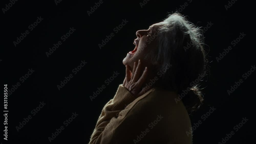 Side studio portrait in profile of senior Caucasian woman in sweater putting hands to head and yelling in pain against black background, feeling hopelessness and despair Side studio portrait in profile of senior Caucasian woman in sweater putting hands to head and yelling in pain against black background, feeling hopelessness and despair