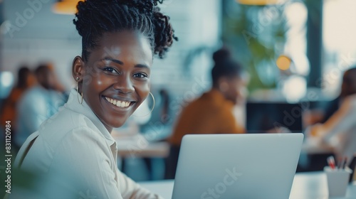 Smiling young African American businesswoman working on a laptop at her desk in a bright modern office with colleagues in the background : Generative AI