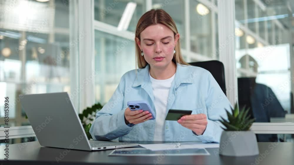 Portrait on serious woman entering card data into personal smartphone at desktop with wireless laptop, documents and decorative plant. Confident caucasian female preparing to pay process indoos. Portrait on serious woman entering card data into personal smartphone at desktop with wireless laptop, documents and decorative plant. Confident caucasian female preparing to pay process indoos.