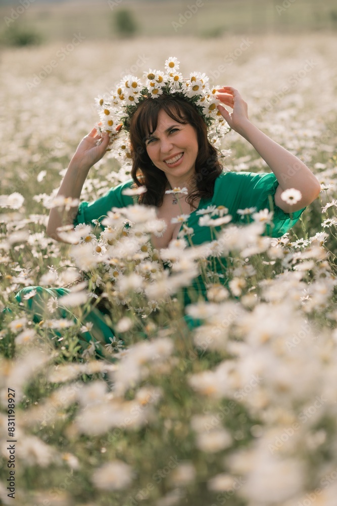 Fototapeta premium Happy woman in a field of daisies with a wreath of wildflowers on her head. woman in a green dress in a field of white flowers. Charming woman with a bouquet of daisies, tender summer photo