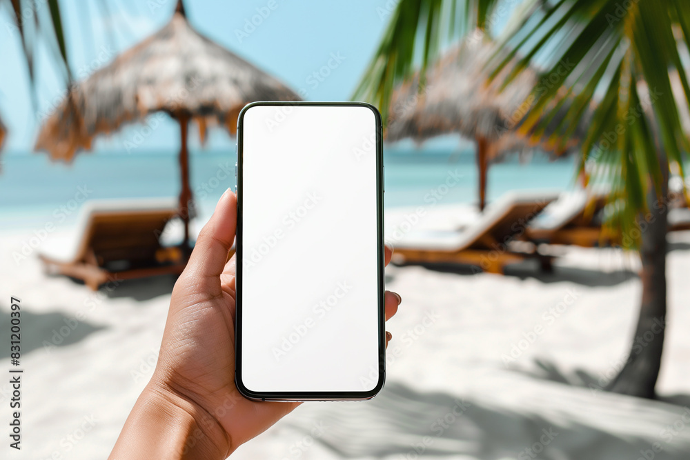 Naklejka premium A close-up shot of a woman's hand holding a blank white screen mobile phone with a tropical beach and sunbeds in the background