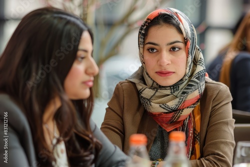 Two diverse women, one wearing a headscarf, are seated at a table with drinks in front of them, engaged in a professional discussion