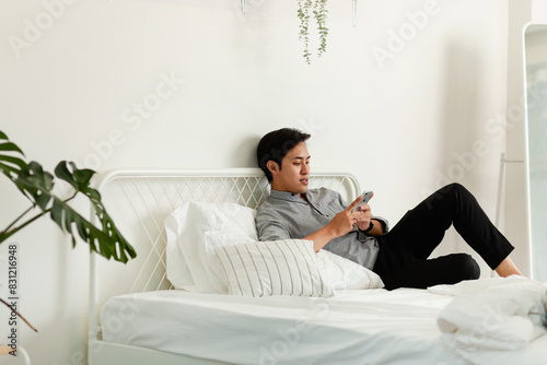 A man is sitting on a bed and looking at his cell phone. The room is clean and well-lit, with a potted plant in the corner. The man is focused on his phone