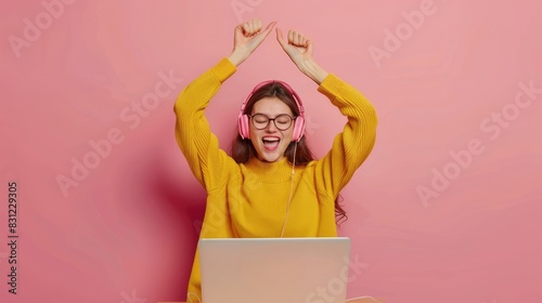 Happy young woman with raised hands sitting in front of laptop with headphones and eyeglasses on isolated pink background with space for copy