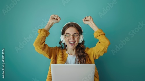 Happy young woman with raised hands sitting in front of laptop with headphones and eyeglasses on isolated green background with space for copy