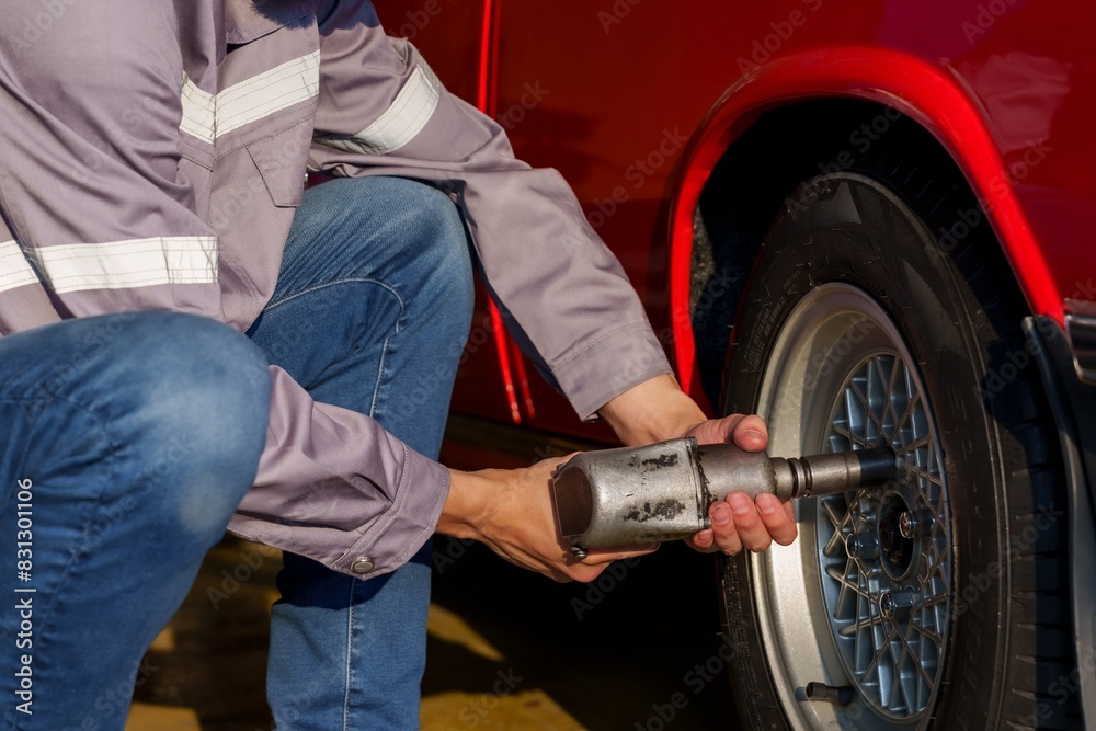Asian male mechanic using power tool on car wheel in auto repair shop. Wearing grey uniform, focusing on precision and task. Scene highlights expertise, dedication, and professional automotive service Asian male mechanic using power tool on car wheel in auto repair shop. Wearing grey uniform, focusing on precision and task. Scene highlights expertise, dedication, and professional automotive service