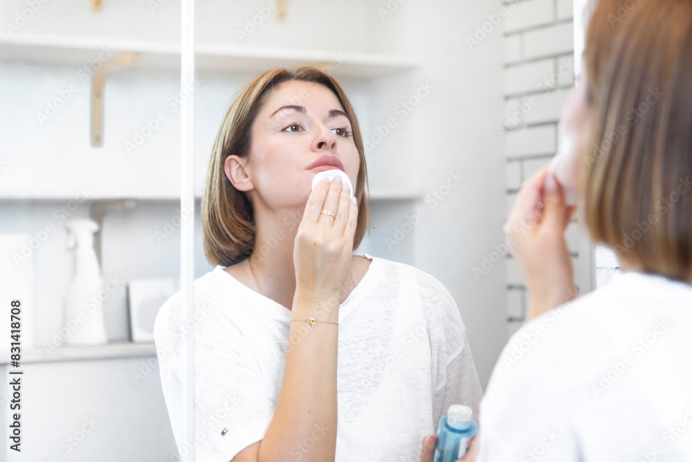 Fototapeta premium Attractive woman looking at mirror and applying moisturizing cream on cheeks in bathroom. Groomed young girl doing skincare morning routine after taking a shower