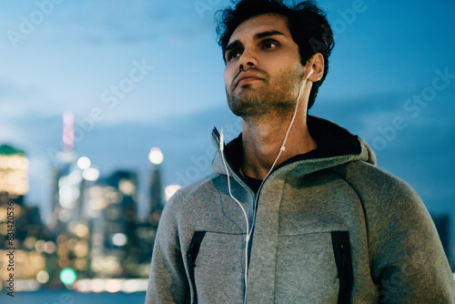 Handsome male meloman dressed in sport wear listening cool music in modern earphones connected to smartphone device standing in urban setting on city promotional background while enjoying evening time photo