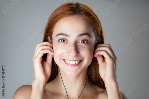 A close-up image of a radiant young woman with red hair, beaming with a joyful smile as she gently holds her earlobes, set against a soft gray backdrop.