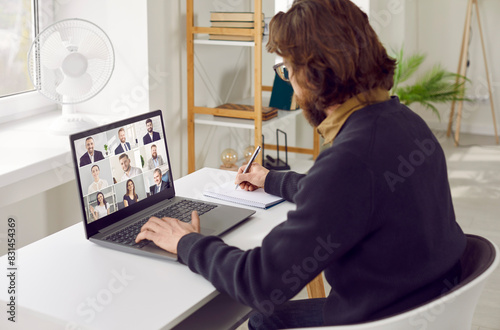 Businessman having online meeting with remote team via conference call. Young male entrepreneur sitting at table with modern laptop computer in homeoffice workplace, attending webinar and taking notes