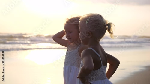 Sisters stand on seashore, watching sunset. Sisters faces with smiles enjoying moment unity with nature. Sisters hold back their hair blowing in strong sea wind. Peaceful atmosphere for communication.