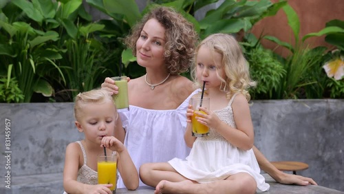 Mom watches with smile as twins happily sip freshly squeezed juice through straws. Happy family happily enjoying juice during summer holidays. Freshly squeezed healthy juice cools down in summer heat.