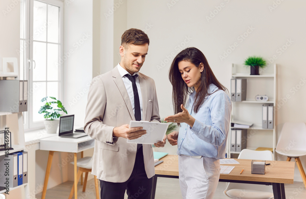 Portrait of two smiling business people man and woman standing in office at their workplace and looking through financial documents. Employees discussing work project or company finances. Portrait of two smiling business people man and woman standing in office at their workplace and looking through financial documents. Employees discussing work project or company finances.