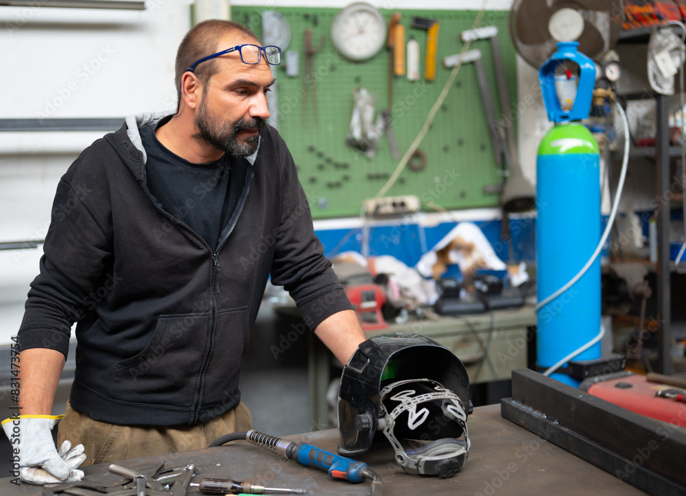 Portrait of a blacksmith during a break in a welding session. He is observing the welding of the two bars on the right. On the workbench the welding helmet and gun. Concept metal craftsmanship,repair. Portrait of a blacksmith during a break in a welding session. He is observing the welding of the two bars on the right. On the workbench the welding helmet and gun. Concept metal craftsmanship,repair.
