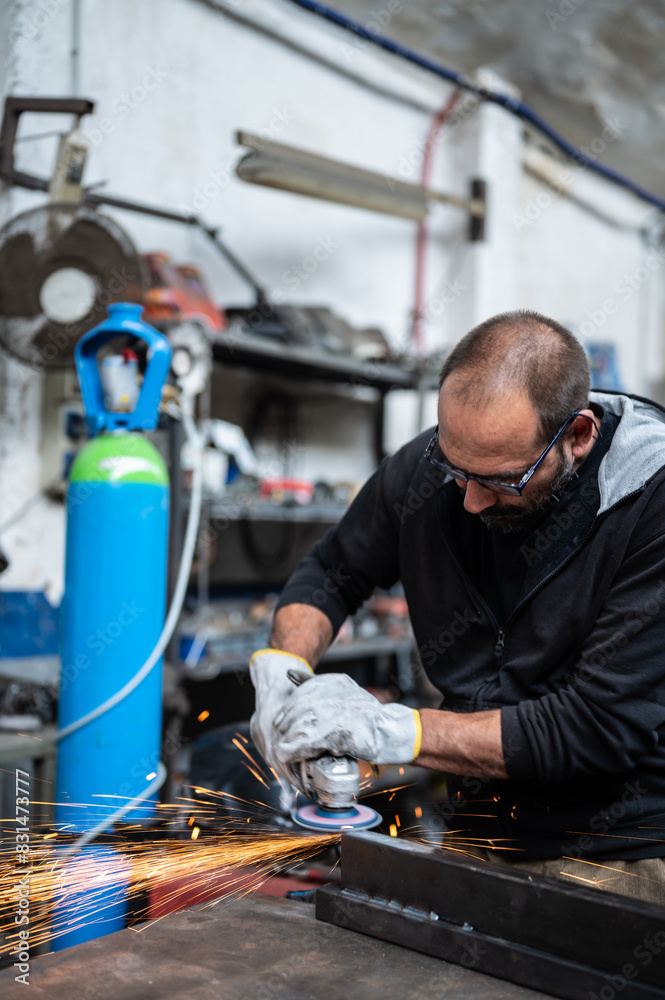 Using the flexible disc grinder on a metal bar: When the tool touches the surface, numerous orange sparks are projected. Vertical format. Concept of metal processing in the workshop, craft work. Using the flexible disc grinder on a metal bar: When the tool touches the surface, numerous orange sparks are projected. Vertical format. Concept of metal processing in the workshop, craft work.