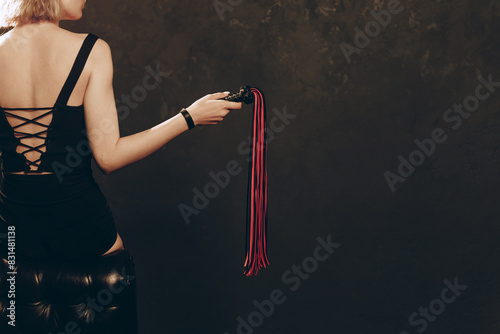 Slender Woman holding Black and Red Leather Flogging Whip in hand in dark interior, view from back