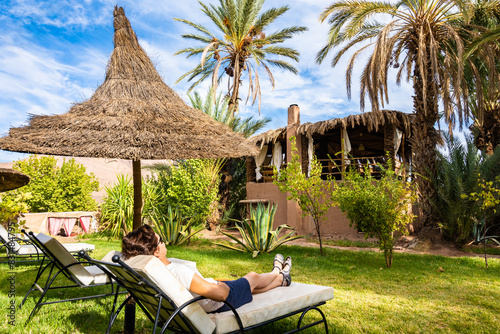 Young woman tourist relaxing on sun chair in tropical gardens of luxury lodge hotel in green palm tree oasis near Agdz town, Morocco, North Africa