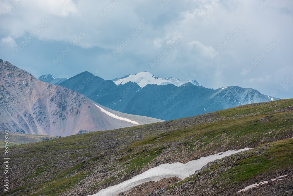 Dramatic layer alpine view to high snowy mountains far away, big rocky ridge, diagonal stony green hillside under rainy gray sky. Large wide snow-capped mountain range silhouette in center close up. Dramatic layer alpine view to high snowy mountains far away, big rocky ridge, diagonal stony green hillside under rainy gray sky. Large wide snow-capped mountain range silhouette in center close up.