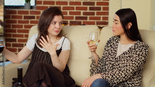 Asian woman chatting with friend on comfortable couch at apartment party, drinking wine and enjoying conversation. Joyful BFFs catching up with each other at home, consuming champagne photo
