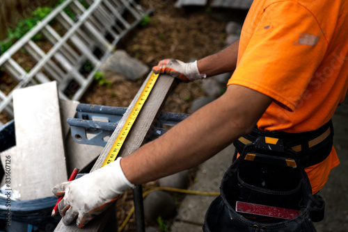 Construction worker in an orange shirt using a tape measure and pencil to mark wood for cutting.