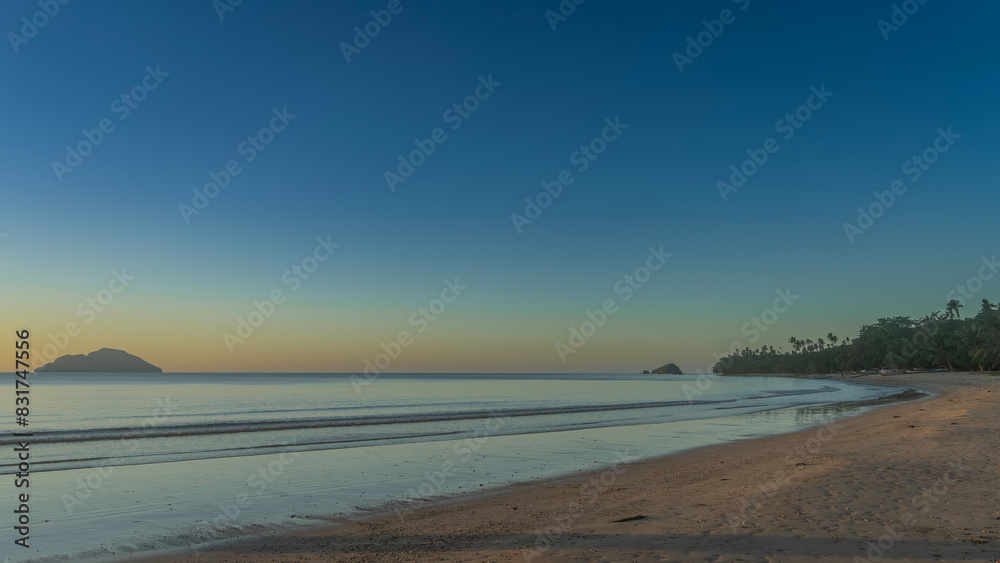 Evening seascape. The waves of the ocean are calmly rolling towards the shore. Shadows on the sandy beach. Green trees, an island in the distance.The blue sky is highlighted with orange at the horizon Evening seascape. The waves of the ocean are calmly rolling towards the shore. Shadows on the sandy beach. Green trees, an island in the distance.The blue sky is highlighted with orange at the horizon