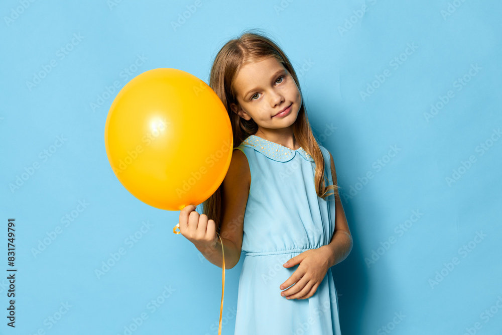 Little girl in blue dress joyfully holding a yellow balloon against a matching blue background Little girl in blue dress joyfully holding a yellow balloon against a matching blue background