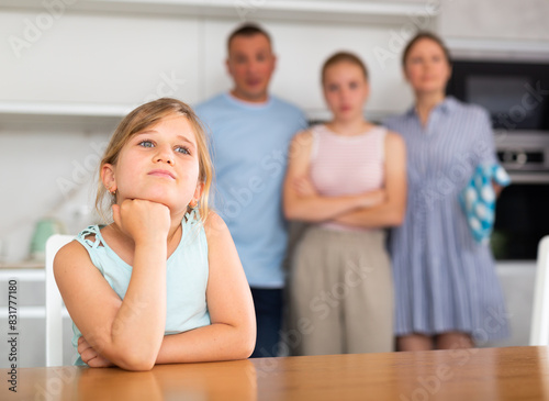 Sad preschooler girl sits at the table while parents and older sister scold © JackF