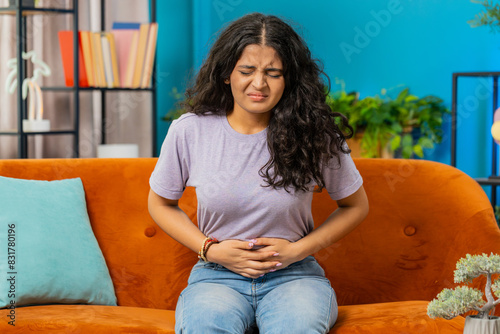 Sick ill Indian woman suffering from period cramps, painful stomach ache on sofa at home room. Girl holding belly, feeling abdominal menstrual pain. Abdominal pain, gastritis, diarrhea, indigestion photo