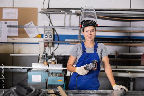 Confident smiling young woman in blue overalls, protective face shield and gloves standing with handheld angle grinder at profile cutting table in window production and assembly workshop..
