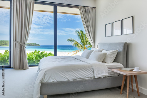 Beautiful contemporary bedroom interior with large windows and a view outside to a tropical beach. Comfortable bed with white bedding and three blank artwork pieces on wall behind.