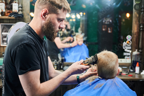Professional barber concentrated on his work. Little kid getting first haircut in barbershop. Male hairdresser using electric shaver to cut boy's hair.